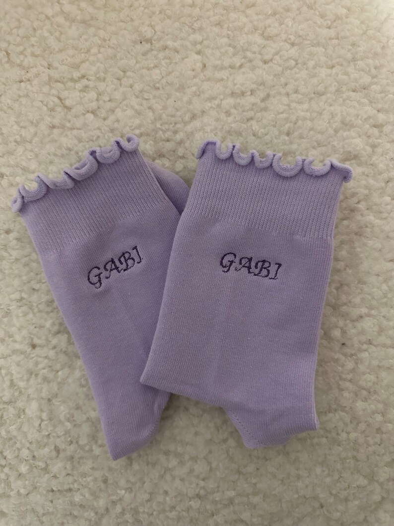 May include: A pair of lavender socks with a frilly top edge. The socks have the name "GABI" embroidered on them.