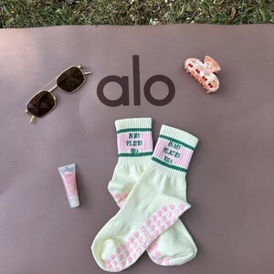 May include: A pair of white socks with pink and green stripes and the text "In My Pilates Era" on the top. The socks have pink heart-shaped grips on the bottom. The socks are on a brown yoga mat with the word "alo" in black letters. There is a pair of sunglasses and a pink hair clip on the mat.