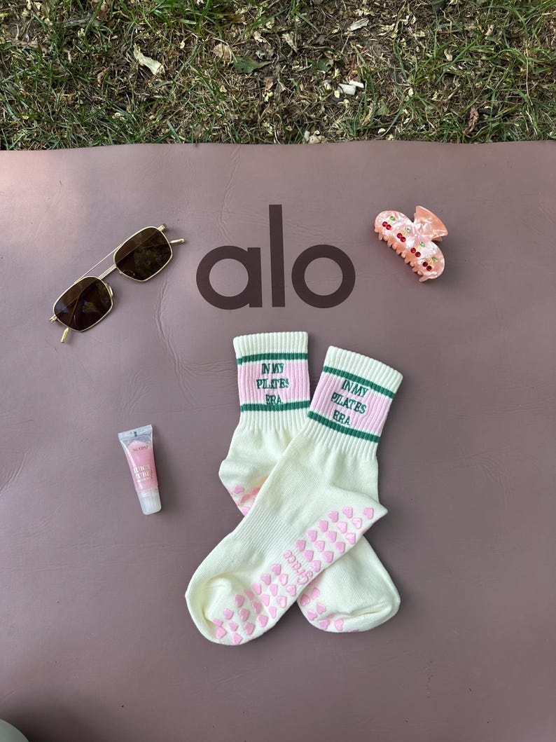May include: A pair of white socks with pink and green stripes and the text "In My Pilates Era" on the top. The socks have pink heart-shaped grips on the bottom. The socks are on a brown yoga mat with the word "alo" in black letters. There is a pair of sunglasses and a pink hair clip on the mat.