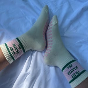 May include: A pair of white socks with pink and green stripes. The socks have the text "IN MY ERA PILATES" on them. The socks have a pattern of pink hearts on the bottom.