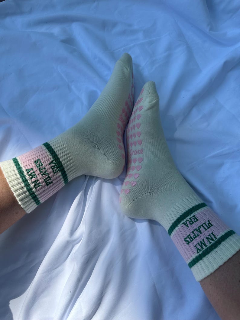 May include: A pair of white socks with pink and green stripes. The socks have the text "IN MY ERA PILATES" on them. The socks have a pattern of pink hearts on the bottom.