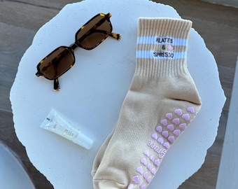 May include: A pair of beige socks with pink heart-shaped grips on the bottom. The socks have white stripes and the text "Pilates & Espresso" is embroidered on the top.
