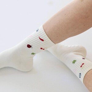 May include: Cream-coloured socks with embroidered patterns. The socks have small, colourful motifs including a ladybird, chilli pepper, and a four-leaf clover. The socks are ribbed and appear to be made of a soft, comfortable material.