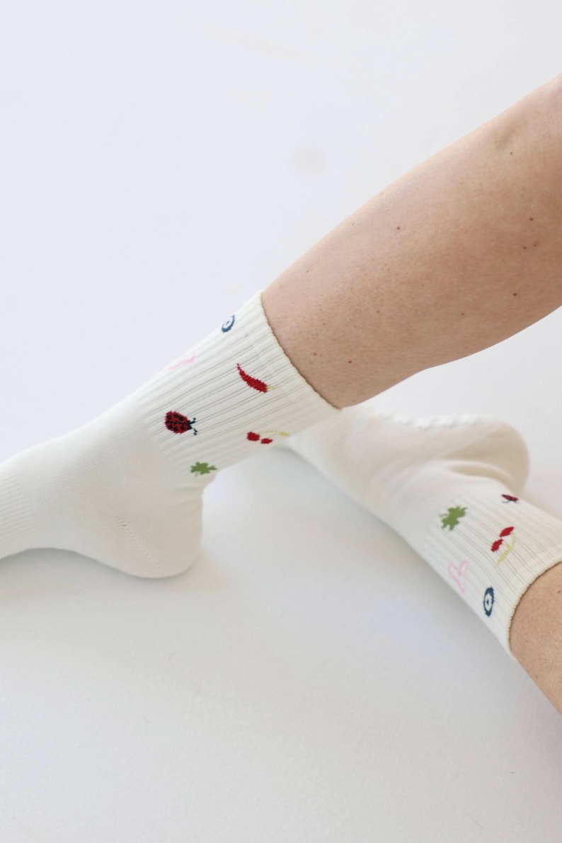 May include: Cream-coloured socks with embroidered patterns. The socks have small, colourful motifs including a ladybird, chilli pepper, and a four-leaf clover. The socks are ribbed and appear to be made of a soft, comfortable material.