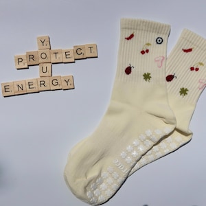 May include: Pair of cream-coloured socks featuring embroidered cherries, clovers, and chillies. The socks have white grip dots on the soles. Wooden letter tiles spell out "PROTECT U ENERGY".