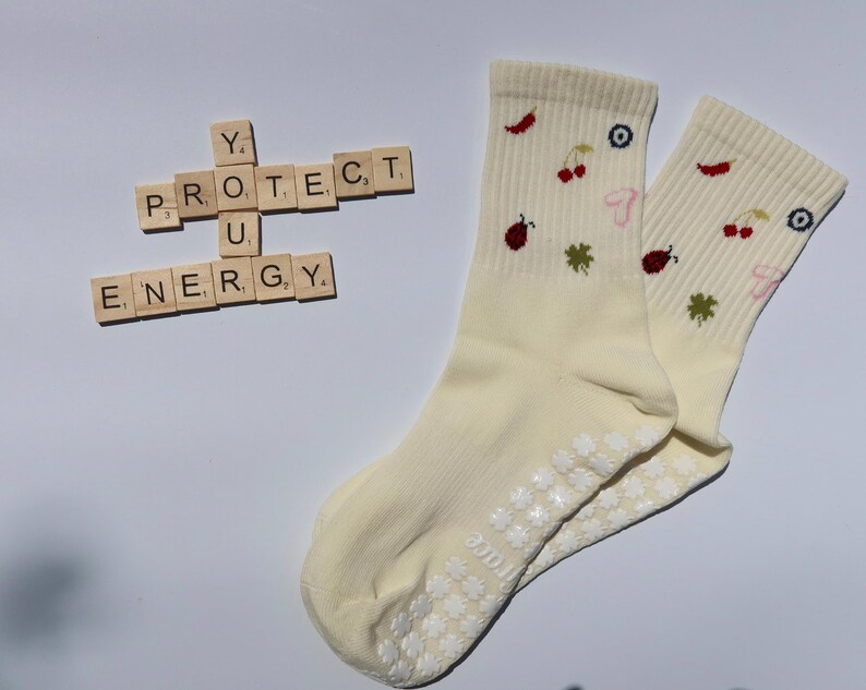 May include: Pair of cream-coloured socks featuring embroidered cherries, clovers, and chillies. The socks have white grip dots on the soles. Wooden letter tiles spell out "PROTECT U ENERGY".