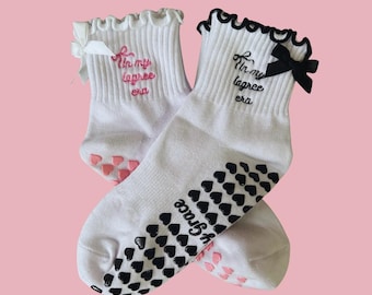 Puede incluir: Un par de calcetines blancos hasta el tobillo con agarres en forma de corazón negros y rosas en las suelas. Los calcetines tienen un ribete de encaje con volantes y un lazo negro en un calcetín. Los calcetines tienen el texto "In my lagree era" bordado.