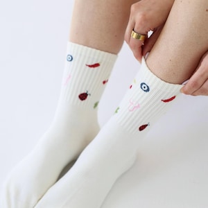 May include: White crew socks with colourful embroidered designs. The socks feature small illustrations of a chilli pepper, a ladybug, an eye, and other shapes. The socks are made of a soft, ribbed material.