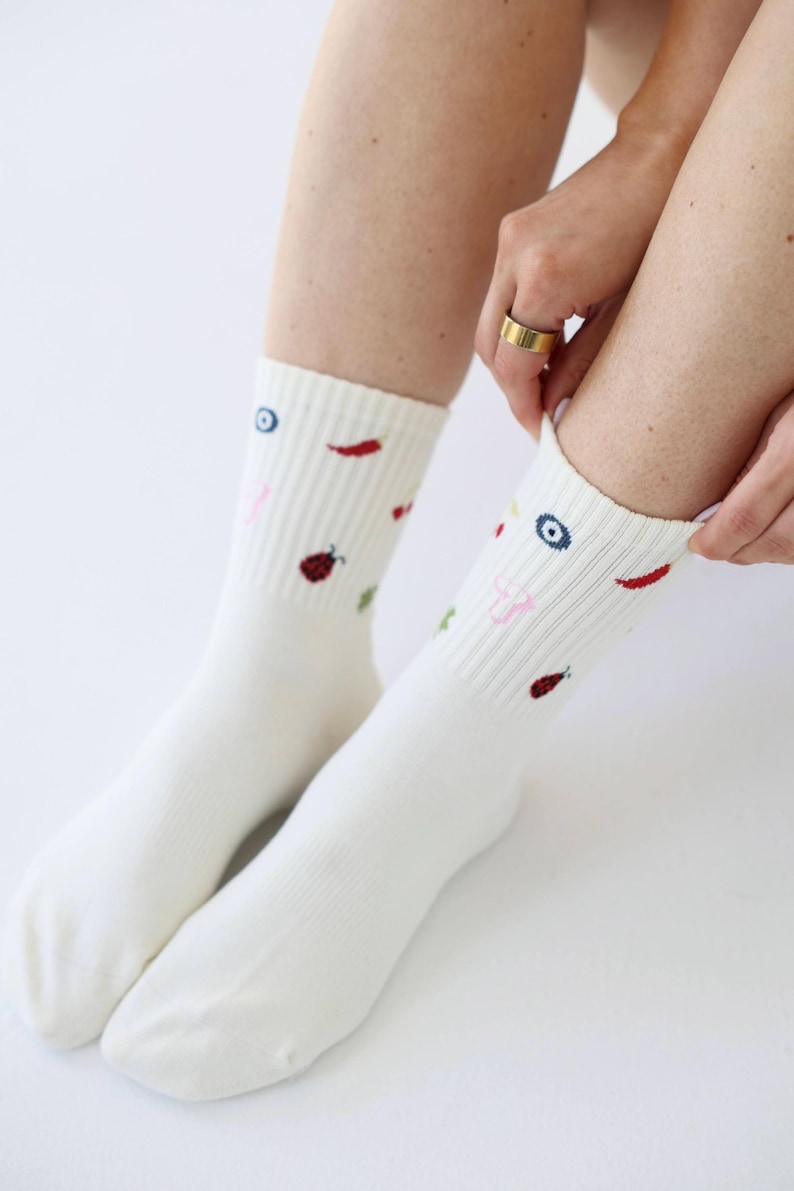 May include: White crew socks with colourful embroidered designs. The socks feature small illustrations of a chilli pepper, a ladybug, an eye, and other shapes. The socks are made of a soft, ribbed material.