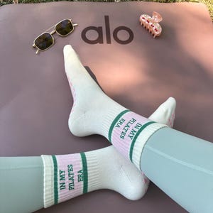 May include: A pair of white socks with pink and green stripes. The socks have the text "IN MY ERA PILATES" written on them.