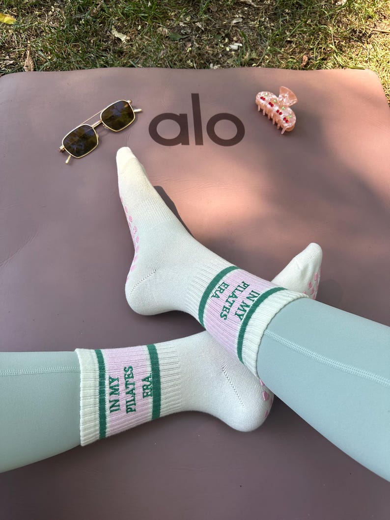 May include: A pair of white socks with pink and green stripes. The socks have the text "IN MY ERA PILATES" written on them.