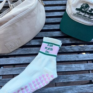May include: A pair of white socks with pink hearts on the bottom and green and pink stripes around the top. The socks say "My Pilates Era" in black text. A beige fanny pack with a zipper and a beige baseball cap with green trim and the text "I Love New York - The World's Borough" are also in the image.