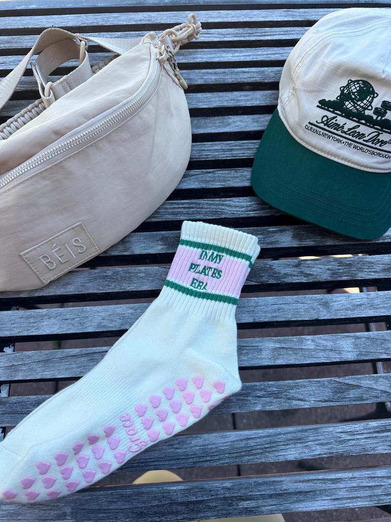 May include: A pair of white socks with pink hearts on the bottom and green and pink stripes around the top. The socks say "My Pilates Era" in black text. A beige fanny pack with a zipper and a beige baseball cap with green trim and the text "I Love New York - The World's Borough" are also in the image.