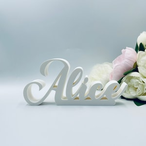Può includere: Un cartello in legno bianco con il nome "Alice" scritto in corsivo. Il cartello è appoggiato su una superficie bianca con un mazzo di fiori bianchi e rosa a destra.