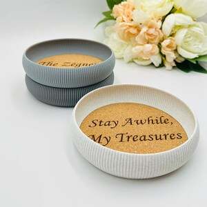 May include: Three round decorative trays with a ribbed exterior. Two are grey, and one is white. Each tray has a cork base with engraved text. The white tray reads "Stay Awhile, My Treasures". A bouquet of flowers is in the background.