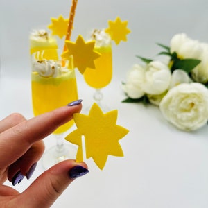 May include: Two yellow cocktails with whipped cream and a yellow star-shaped garnish. A hand holds a yellow star-shaped garnish. A straw is in one of the cocktails. White flowers are in the background.