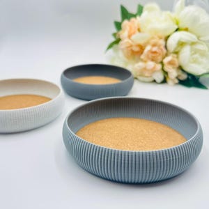 May include: Three decorative bowls in shades of gray and white, each with a cork base. The bowls have a ribbed exterior design. A bouquet of flowers is in the background. Home decor.
