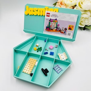 May include: A light teal, pentagonal plastic sorting tray with six compartments containing various small building bricks and a LEGO Gabby's Dollhouse 10795 instruction manual. The name Jasmina is displayed in yellow 3D letters on the back panel.