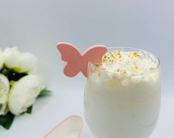 May include: A glass of white milk topped with whipped cream and gold sprinkles, garnished with a pink butterfly-shaped decoration.