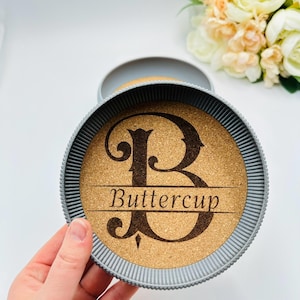 May include: A round cork coaster with a grey rim, featuring a large letter "B" and the name "Buttercup" engraved in a decorative font. The coaster is held in a grey holder.