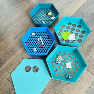 May include: A collection of five hexagonal, plastic storage trays in shades of teal and blue. The trays have perforated or circular designs and contain small toy building blocks. One tray has a solid teal lid with two circular cutouts. The trays are on a wooden floor.