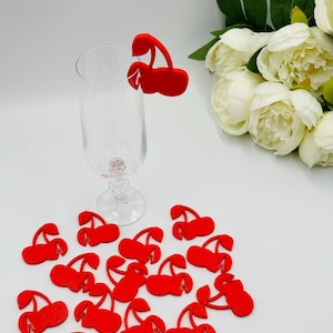 May include: Red cherry shaped glass markers, scattered on a white surface. One cherry is on a clear glass.