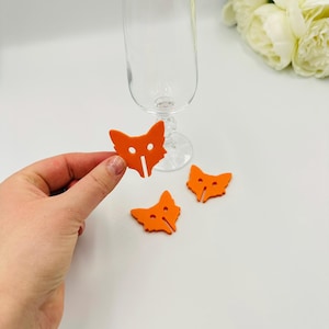 May include: Three orange fox-shaped glass markers. The markers have a cut-out design of a fox's face with a black nose and eyes.