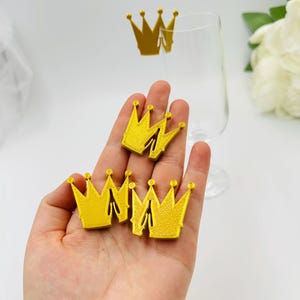 May include: Four gold crown shaped paper drink charms. The charms are in the palm of a hand.