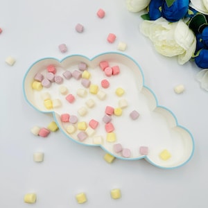 Puede incluir: Una fuente en forma de nube con borde azul claro, llena de caramelos cuadrados de colores en tonos pastel de rosa, amarillo y lavanda. Algunos caramelos tienen diseños florales en relieve. Los caramelos están esparcidos alrededor de la fuente sobre una superficie blanca.