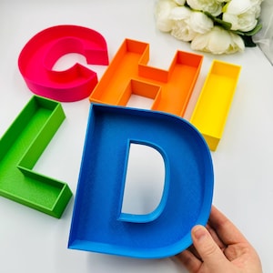 Pode incluir: Letras 3D coloridas que soletram "CHILD". As letras são em cores vibrantes: rosa, laranja, verde, azul e amarelo. As letras são feitas de um material espesso e têm formato de caixa. A letra "D" está sendo segurada por uma mão.