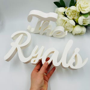 Puede incluir: Un letrero de madera blanco con el texto "The Ryals" en una fuente cursiva. El letrero está apoyado en una superficie blanca.