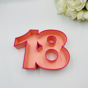 May include: A pink and fuchsia number 18 cake topper. The number is made of a thick, sturdy material and has a hollow center.