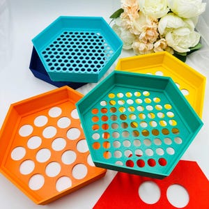 May include: A collection of colorful, hexagonal trays in shades of blue, orange, yellow, green, and red. Each tray features a unique design, with some having perforated holes and others with circular cutouts. The trays are arranged on a white surface.