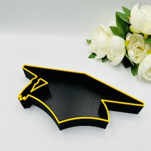 May include: A black tray in the shape of a graduation cap, with a yellow outline. The tray has a textured surface. White flowers are visible in the background.