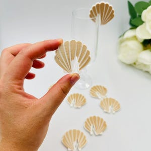 Op de afbeelding: Schelpvormige drankbedels in beige en wit. Eén is bevestigd aan een helder glas, andere liggen op een wit oppervlak. De bedels hebben een gestructureerd, waaiervormig ontwerp. Perfect voor feesten.