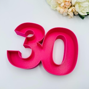 Op de afbeelding: Een felroze nummer 30, waarschijnlijk voor een verjaardag of jubileum. De cijfers zijn driedimensionaal en staan rechtop. Op de achtergrond is een boeket witte en perzikroze bloemen te zien.
