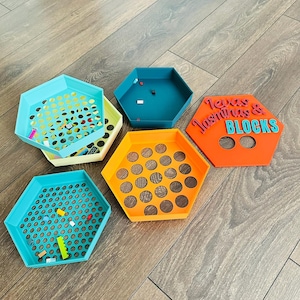 May include: A collection of colorful, hexagonal trays in various shades of blue, orange, and yellow. Some trays have perforated designs, while others are solid. One tray features the text "Texas & Jasmina's BLOCKS". The trays are designed to hold small building blocks.