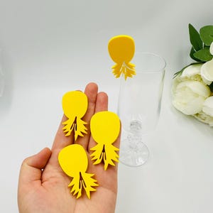 May include: Bright yellow drink charms in a pineapple shape. Three charms are held in a hand, and one is attached to a clear glass. The charms have a rounded top and a detailed, spiky base. The background is white.