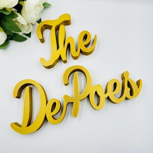 Op de afbeelding: Goudkleurig houten bord met de woorden "The DePoes" in een sierlijk lettertype. Het bord bestaat uit twee afzonderlijke stukken, met "The" bovenaan en "DePoes" eronder. Geschikt voor woondecoratie of als gepersonaliseerd cadeau.