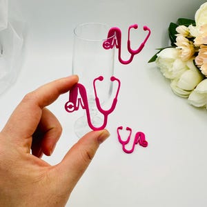Peut inclure: Charmant porte-verres en forme de stéthoscope rose vif. Plusieurs breloques sont dispersées autour d'une flûte à champagne, certaines étant tenues dans une main. Les breloques sont d'un rose fuchsia vif, parfaites pour un événement médical ou à thème.