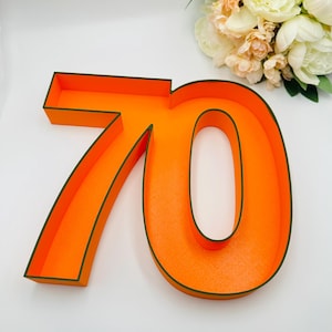 May include: Vibrant orange number 70 with a dark green trim, likely for a celebration. The number has a three-dimensional design. A bouquet of white and peach flowers is in the background, adding a decorative touch.