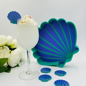 Op de afbeelding: Een set van vijf teal en paarse drinkonderzetters in de vorm van een schelp met een grotere teal en paarse schelpvormige dienblad. De onderzetters en het dienblad hebben een reliëf, textuurontwerp.