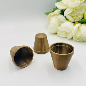 Pode incluir: Três pequenos vasos de flores marrons vazios de várias formas são dispostos sobre uma superfície branca. Os vasos são feitos de um material fosco. Flores brancas com folhas verdes ao fundo.