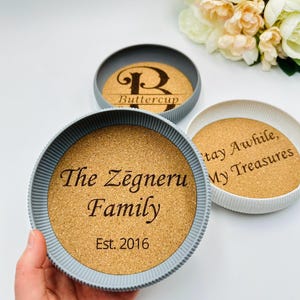Può includere: Tre sottobicchieri rotondi con centri in sughero e bordi grigi o bianchi. Un sottobicchiere recita "The Zēgneru Family Est. 2016", un altro dice "Buttercup" e il terzo dice "Stay Awhile, My Treasures".