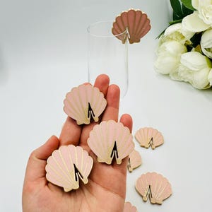 May include: Pink and gold seashell-shaped drink charms. The charms are made of wood and have a slot to attach to a glass. One charm is attached to a clear glass. The charms are a light pink color with gold accents.
