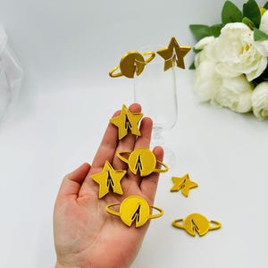 Può includere: Un set di ciondoli per bevande in carta tagliati al laser color oro, raffiguranti una stella e un pianeta con anelli. I ciondoli sono perfetti per aggiungere un tocco di fantasia a qualsiasi festa o evento.