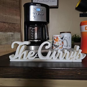 Può includere: Un cartello in legno bianco con il testo "The Curry's" in un font corsivo. Il cartello è appoggiato su una superficie in legno scuro davanti a una macchina per il caffè e due tazze.
