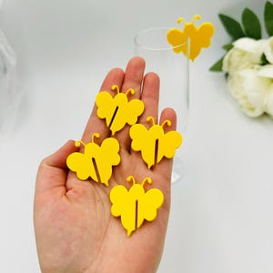 Könnte beinhalten: Fünf gelbe, schmetterlingsförmige Glasmarker. Die Marker sind schmetterlingsförmig mit einem Ausschnitt in der Mitte für ein Glas.