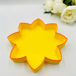 Puede incluir: Una bandeja amarilla con forma de flor con un borde naranja brillante. La bandeja está sobre un fondo blanco, con un ramo borroso de flores blancas en el fondo. El diseño de la bandeja es simple y moderno.