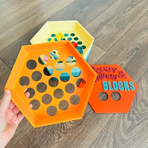 May include: Three hexagonal trays in yellow, orange, and blue. The orange tray has multiple circular cutouts. The yellow tray contains small, colorful toy blocks. The orange tray has the text "Texas Mines & Blocks" in blue.